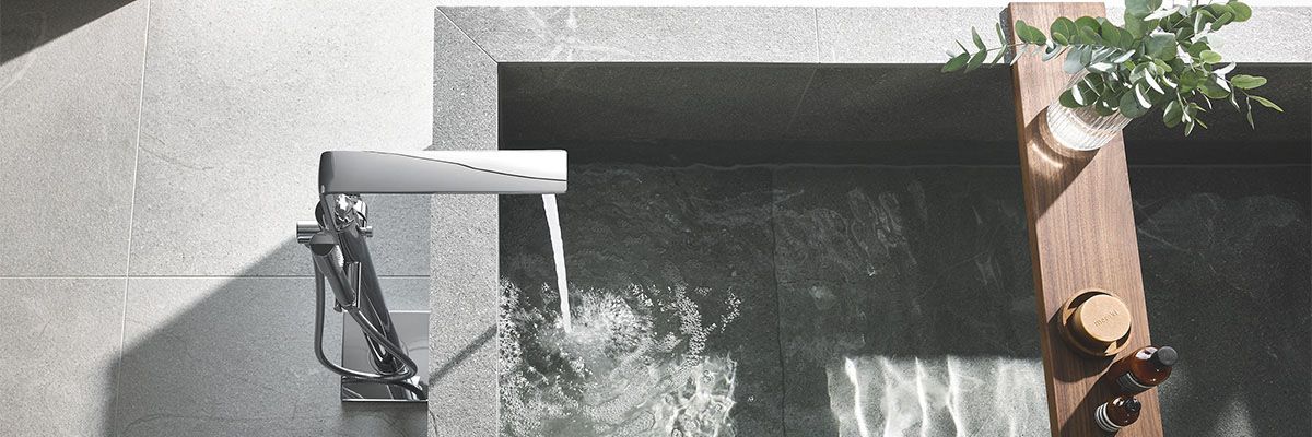 Eine Steinbadewanne mit einer Badewannenbrücke mit einigen Gegenständen darauf von oben. Der Wasserhahn ist an und lässt Wasser ins Bad ein.